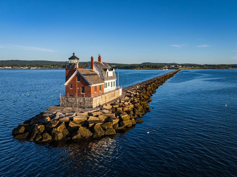 Scenic view of a historic lighthouse on a rocky breakwater extending into the calm blue sea under a clear sky -cm